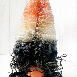 May include: A small orange, white, and black bottle brush tree gnome with a black ribbon and a black pot. The tree is covered in white faux snow.