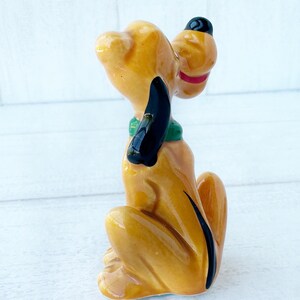 May include: A ceramic figurine of a seated dog with a yellow body, black ears, and a black hat. The dog has a green collar and a red mouth. The figurine is a collectible item.