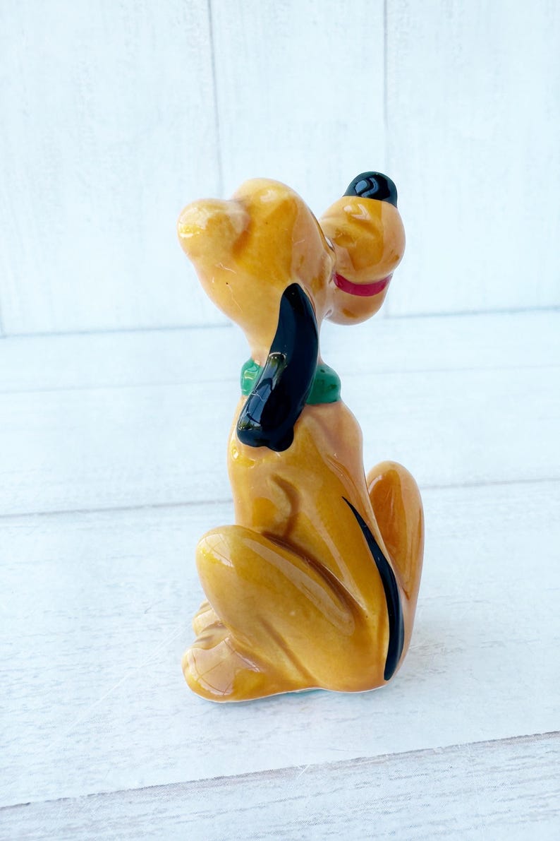 May include: A ceramic figurine of a seated dog with a yellow body, black ears, and a black hat. The dog has a green collar and a red mouth. The figurine is a collectible item.