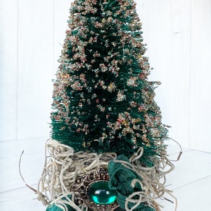 May include: A small green bottle brush Christmas tree decorated with gold and red glitter, nestled in a bed of natural fiber and accented with a green ribbon and a large, ornate, jeweled brooch.