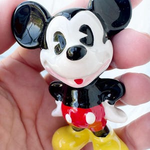 May include: A small ceramic Mickey Mouse figurine. The figure has black ears, a black body, white face, red shorts with white buttons, and yellow shoes. The figurine is held in a person's hand.