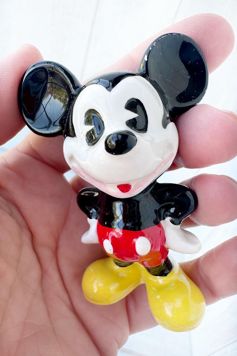 May include: A small ceramic Mickey Mouse figurine. The figure has black ears, a black body, white face, red shorts with white buttons, and yellow shoes. The figurine is held in a person's hand.