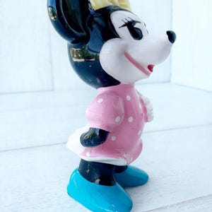 May include: A ceramic Minnie Mouse figurine. It has a black head and ears, a yellow bow, and a pink dress with white polka dots. She wears blue shoes and a white skirt. A collectible item.