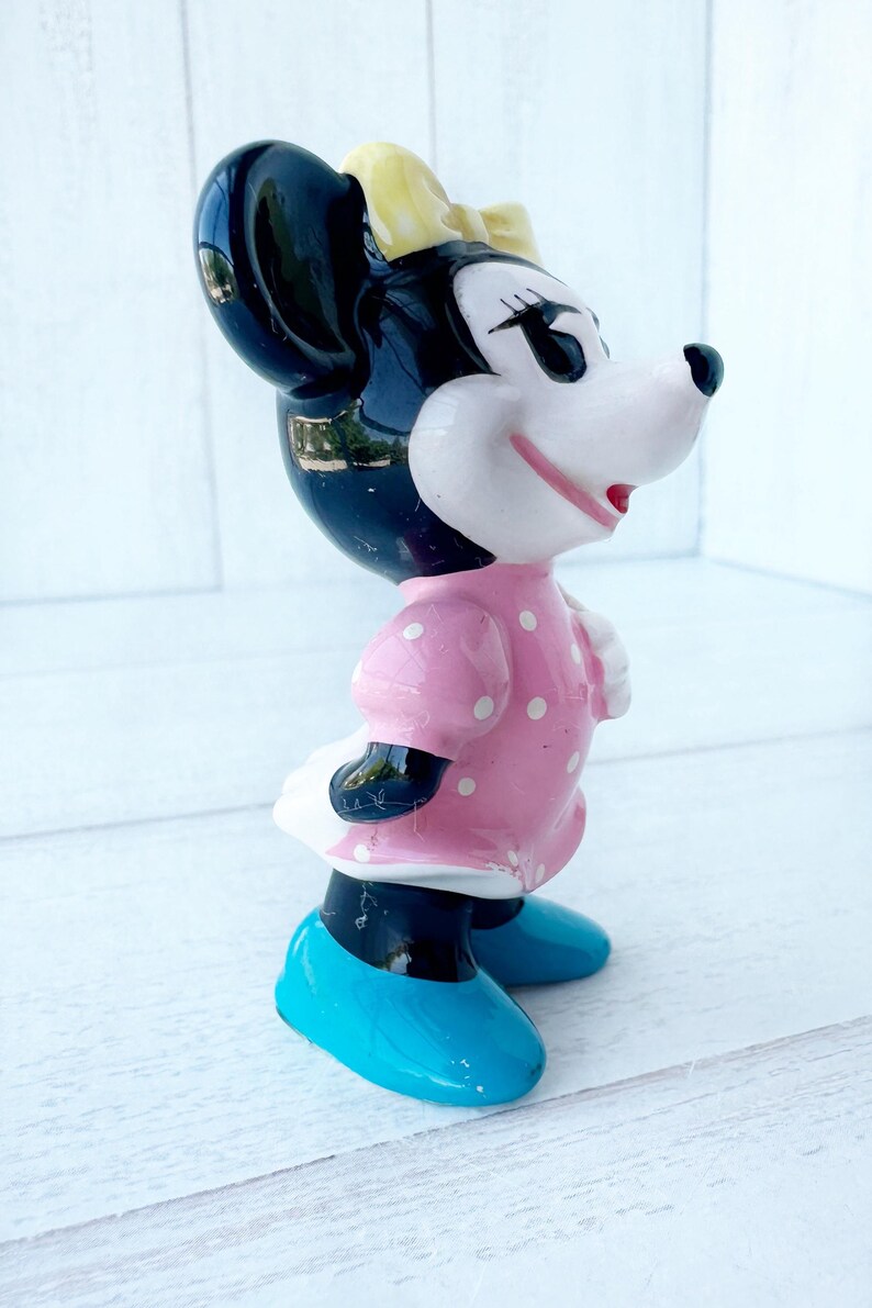 May include: A ceramic Minnie Mouse figurine. It has a black head and ears, a yellow bow, and a pink dress with white polka dots. She wears blue shoes and a white skirt. A collectible item.