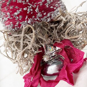 May include: A silver bell with a pink ribbon and a sparkly jewel on top. The bell is surrounded by dried moss.