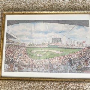 May include: A framed watercolor print of a baseball stadium filled with spectators. The artwork depicts a game in progress, with players on the field and fans in the stands. The frame is gold-colored.