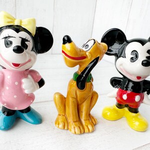 May include: Three ceramic Disney character figurines: Minnie Mouse in a pink dress, Pluto the dog, and Mickey Mouse in red shorts. The figurines are brightly coloured and stand on a white surface.