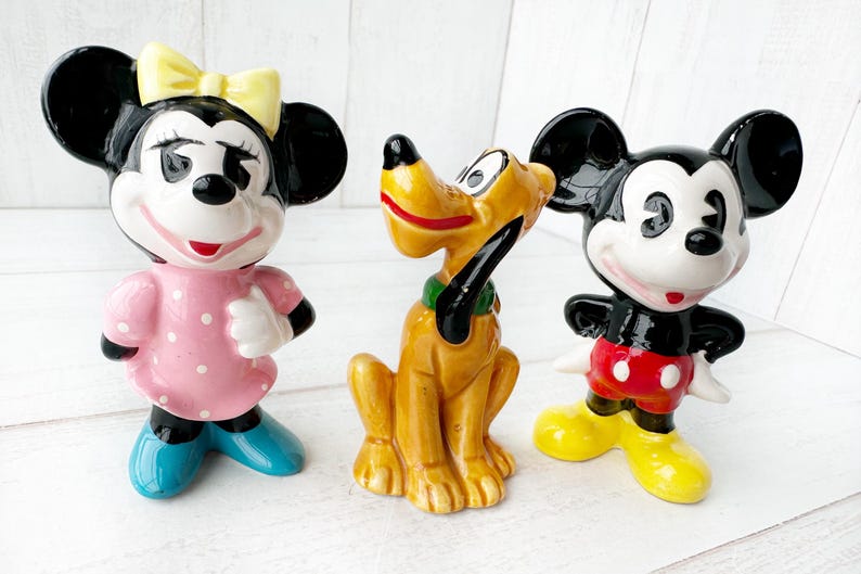 May include: Three ceramic Disney character figurines: Minnie Mouse in a pink dress, Pluto the dog, and Mickey Mouse in red shorts. The figurines are brightly coloured and stand on a white surface.