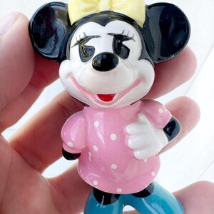 May include: A ceramic figurine of a cartoon mouse character. The mouse has black ears, a yellow bow, and a pink dress with white polka dots. The mouse is wearing blue shoes and has a smiling expression.