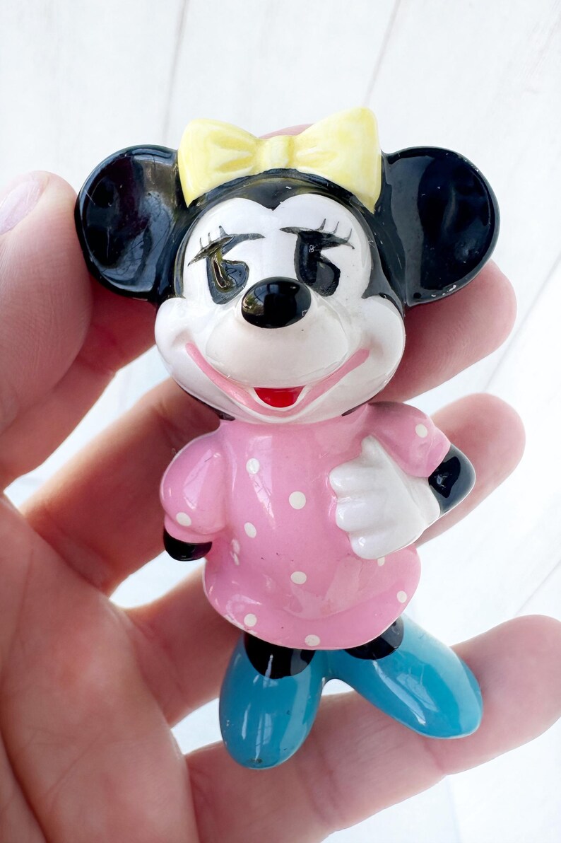 May include: A ceramic figurine of a cartoon mouse character. The mouse has black ears, a yellow bow, and a pink dress with white polka dots. The mouse is wearing blue shoes and has a smiling expression.