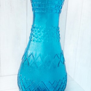 May include: A turquoise blue glass vase with a textured, embossed design. The vase has a wide mouth and a narrow neck.