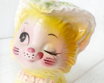 May include: A yellow ceramic piggy bank shaped like a cat's head with a winking eye, pink cheeks, and a tongue sticking out. The base of the bank is yellow with a pink bow and the word "Here" written on it.