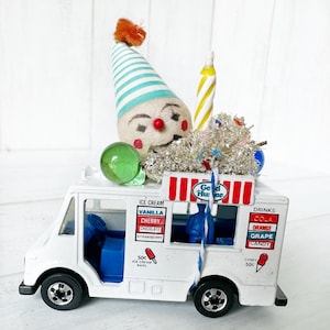 May include: A white toy ice cream truck with a red and white striped awning. The truck has a clown face with a red nose and blue eyes on top. The truck has a sign that says "Good Humor" and has a variety of ice cream and drink options listed on the sides.