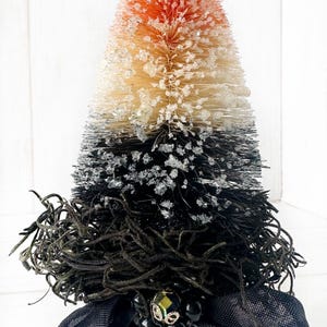 May include: A black and orange bottle brush tree with a black base and black ribbon. The tree is decorated with black and green beads and a gold bead.