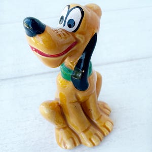 May include: A ceramic Pluto figurine. The cartoon dog is yellow with a black nose and ears, a red mouth, and a green collar. The dog is sitting with its legs tucked under. A collectible item.