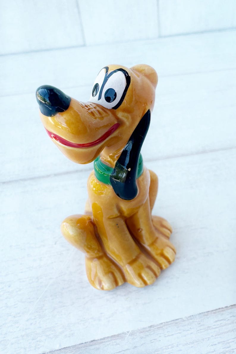 May include: A ceramic Pluto figurine. The cartoon dog is yellow with a black nose and ears, a red mouth, and a green collar. The dog is sitting with its legs tucked under. A collectible item.