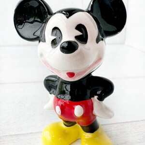 May include: A ceramic Mickey Mouse figurine with a glossy finish. The figure has black ears, a black torso, red shorts with white buttons, yellow shoes, and a white face. The figurine is posed with arms at its sides.