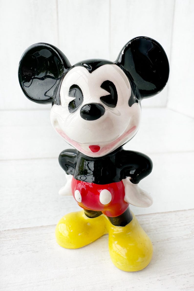 May include: A ceramic Mickey Mouse figurine with a glossy finish. The figure has black ears, a black torso, red shorts with white buttons, yellow shoes, and a white face. The figurine is posed with arms at its sides.