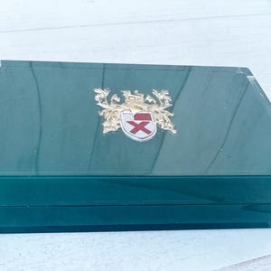 May include: A rectangular, emerald green box with a clear, beveled lid. A gold-colored emblem with a red cross and crown is centered on the lid. The box is likely for jewelry or small keepsakes.