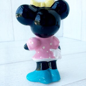 May include: A ceramic mouse figurine with a black head, large ears, and a yellow bow. It wears a pink dress with white spots, white gloves, and blue shoes. The figurine is standing on a white surface.