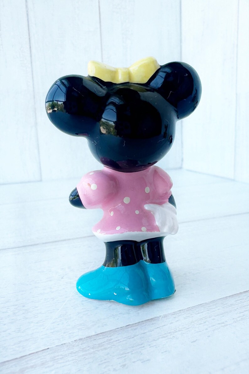 May include: A ceramic mouse figurine with a black head, large ears, and a yellow bow. It wears a pink dress with white spots, white gloves, and blue shoes. The figurine is standing on a white surface.