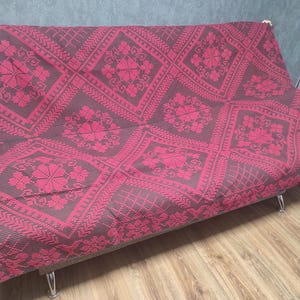 May include: A red and dark brown patterned sofa cover. The cover features a repeating floral and geometric design. The sofa has a gray fabric base and silver legs. The cover is draped over the sofa.