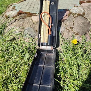 Puede incluir: Un termómetro negro con una escala de temperatura beige y un tubo naranja. El termómetro está en un estuche negro abierto, apoyado sobre una superficie de piedra con hierba verde en el fondo. La escala de temperatura llega hasta los 250 grados.