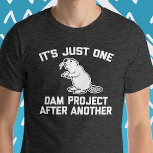 May include: A dark heather grey t-shirt with a white graphic of a beaver holding a branch in its mouth. The text "It's just one dam project after another" is printed below the beaver.