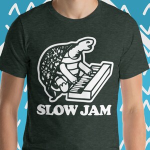 May include: A dark green t-shirt with a white graphic of a turtle playing a keyboard. The text "SLOW JAM" is printed below the graphic.