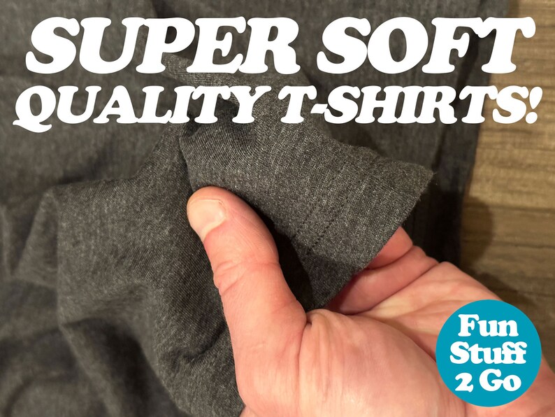 May include: Close-up of a dark gray t-shirt being held in a hand, showing the soft fabric texture. The text "SUPER SOFT QUALITY T-SHIRTS!" is visible at the top of the image. A blue circle with the text "Fun Stuff 2 Go" is visible in the bottom right corner.
