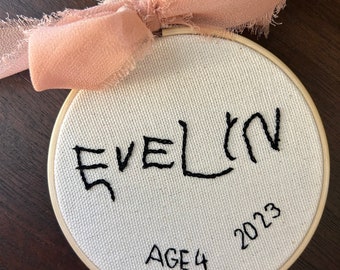 May include: A round, off-white fabric embroidery hoop with the name "EVELIN" stitched in black thread. Below the name are the words "AGE 4" and the year "2023". A dusty rose ribbon is tied at the top.