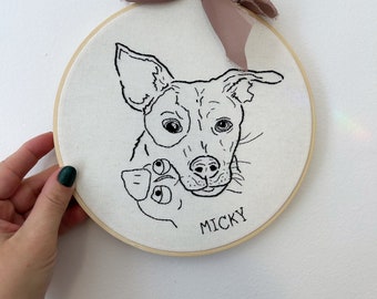 May include: An embroidered hoop art piece featuring a black and white line drawing of a dog's face, with the name "MICKY" below. The artwork is mounted in a wooden hoop and adorned with a mauve ribbon.
