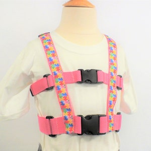 May include: Pink safety harness with adjustable straps and a puzzle piece pattern. The harness has two black buckles.