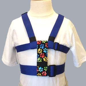 May include: A blue adjustable harness with a colorful paw print pattern on the chest piece. The harness is designed for children and has adjustable straps.