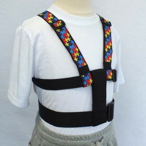 May include: An adjustable harness with puzzle piece patterned straps. The harness is black and blue, with a buckle closure, and is designed for children.