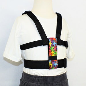 May include: A black adjustable harness with a colorful puzzle piece pattern fabric strap. The harness has two shoulder straps, a chest strap, and a waist strap.