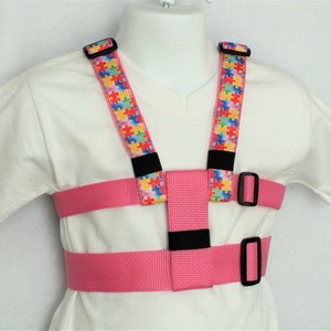 May include: Pink safety harness with adjustable straps and black buckles. The straps have a colorful puzzle piece pattern.