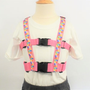 May include: Pink safety harness with adjustable straps and black buckles. The straps are decorated with a colorful puzzle piece pattern.