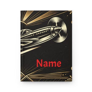 May include: Black journal with a gold art deco design featuring an airplane and the word "Name" in red. The cover has a gold sunburst design and a gold border.