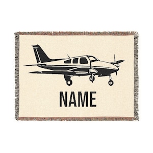 Op de afbeelding: Crèmekleurige geweven sprei met een zwarte vliegtuigillustratie en het woord "NAME" in vette, zwarte letters. De deken heeft een gefranjerde rand.