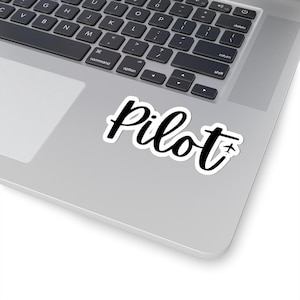 May include: A white sticker with the word "Pilot" in black cursive script. A small airplane graphic is next to the last letter. The sticker is on a silver laptop.