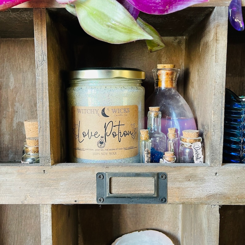 Op de afbeelding: Een houten plank toont een verzameling flessen in apothekersstijl en een grote kaars. De kaars, met het opschrift "Love Potion", heeft een gouden deksel en cr&egrave;me-kleurig was. Er zijn ook verschillende kleine glazen flesjes met kurken te zien.