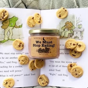 May include: A brown soy wax candle with a white label that reads "Witchy Wicks We Must Stop Eating! 100% Soy Wax". The candle is surrounded by cookies on a book with text that reads "We must stop eating!" and "You mean like trying not to eat all of these asked Toad."
