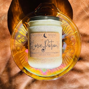 Op de afbeelding: Een glazen potkaars met een zilveren deksel, met het label "Love Potion" op een bruin etiket. De kaars staat in een decoratieve, iriserende glazen schaal. De kaars heeft een gespikkelde uitstraling en de tekst "100% SOY WAX".