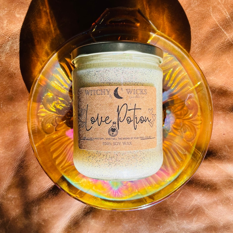 Op de afbeelding: Een glazen potkaars met een zilveren deksel, met het label "Love Potion" op een bruin etiket. De kaars staat in een decoratieve, iriserende glazen schaal. De kaars heeft een gespikkelde uitstraling en de tekst "100% SOY WAX".