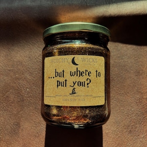 May include: A brown glass jar candle with a gold glitter top and a brown paper label that reads "WITCHY WICKS ...but where to put you? BRAVERY LOYALTY INTELLIGENCE OR AMETION 100% SOY WAX".