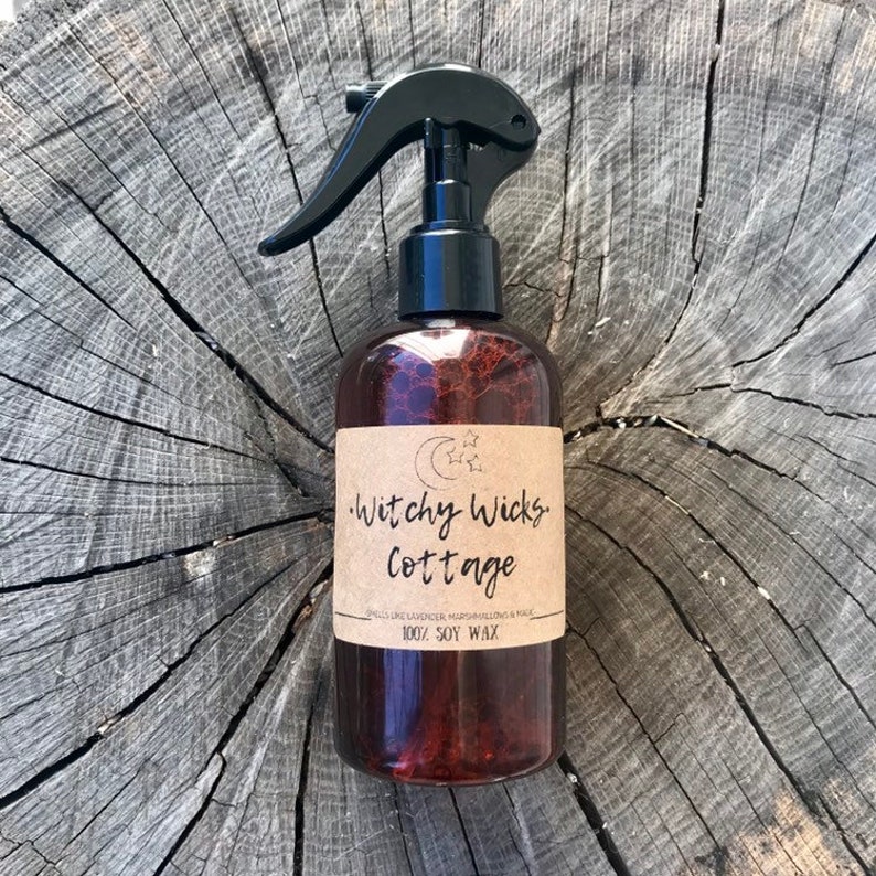 Op de afbeelding: Een bruine spuitfles met een zwarte sproeikop en een etiket met de tekst "Witchy Wicks Cottage". Het etiket bevat ook de tekst "Smells like Lavender, Marshmallow & Musk" en "100% Soy Wax".