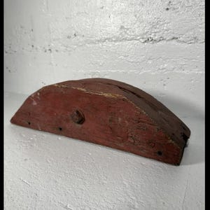 May include: A weathered, semi-circular wooden object painted in a faded red hue. A single, rusty bolt secures a piece of the wood. The object has a rustic, antique appearance, suggesting it may be a decorative or functional piece.