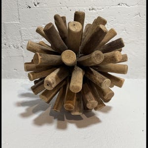 May include: A decorative sphere made of numerous small, natural-toned wooden dowels radiating outwards. The wooden dowels are various shades of brown and tan, creating a textured, spiky appearance. The sphere sits on a white surface.