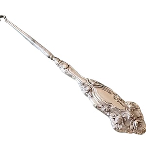 May include: A silver crochet hook with a decorative handle. The hook has a long, slender shaft and a curved end. The handle is ornate, with floral and scroll designs. The word "STERLING" is engraved on the handle.